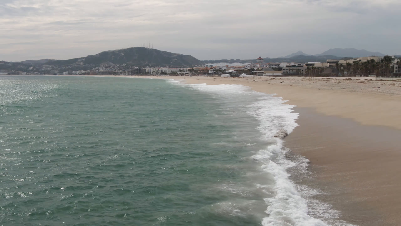 Aerial view of surf washing along a wide beach with hills beyond town buildings.
