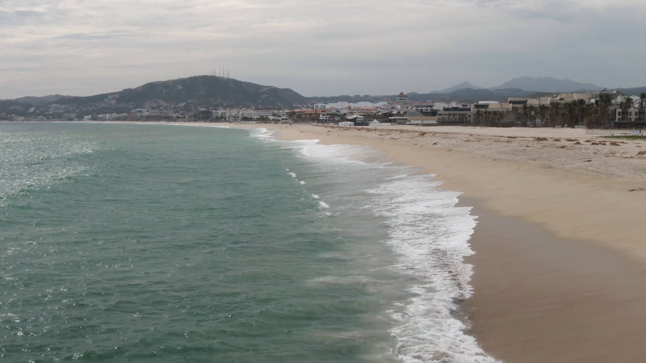 Aerial shoreline view in San Jose del Cabo showing a diagonal band of white surf, green-blue water, broad sand, low buildings near the beach, hazy hills, and no clearly visible people.