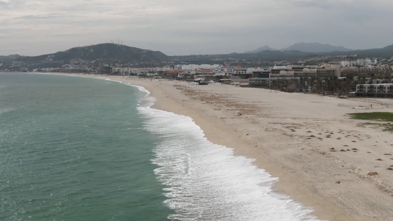 Aerial beach shoreline with ocean, sand, buildings, and distant hills.