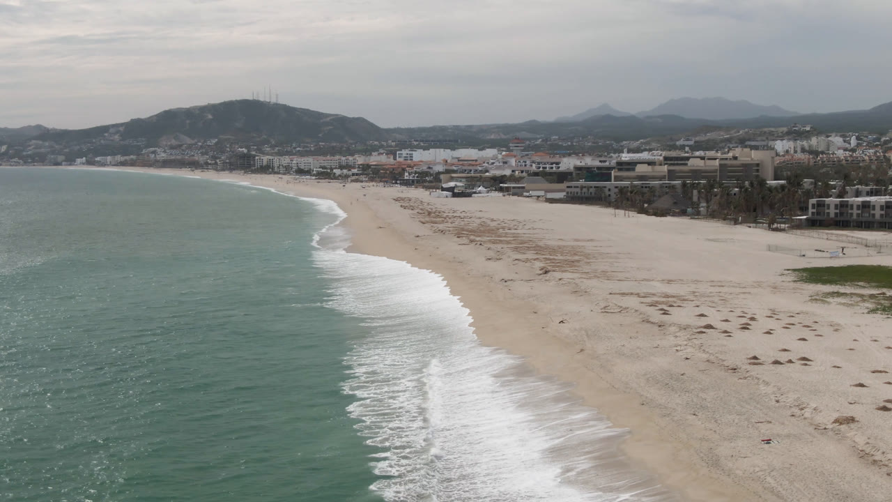 Elevated San Jose del Cabo coastline view one second in, showing a longer sweep of white surf against pale sand, green-blue water, low buildings, hills, and tiny distant beach figures.