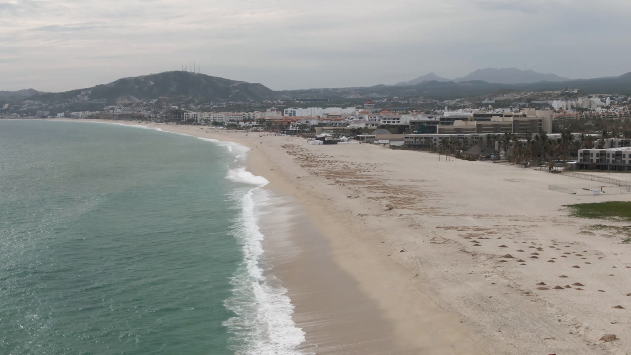 Opening aerial frame over San Jose del Cabo with turquoise water along the left edge, a wide sandy beach curving toward low buildings and hills, and no visible people.