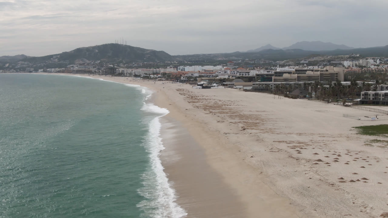 San Jose del Cabo shoreline from above with the wave line running diagonally up the beach, low urban development beyond the sand, hazy hills behind it, and no visible people.