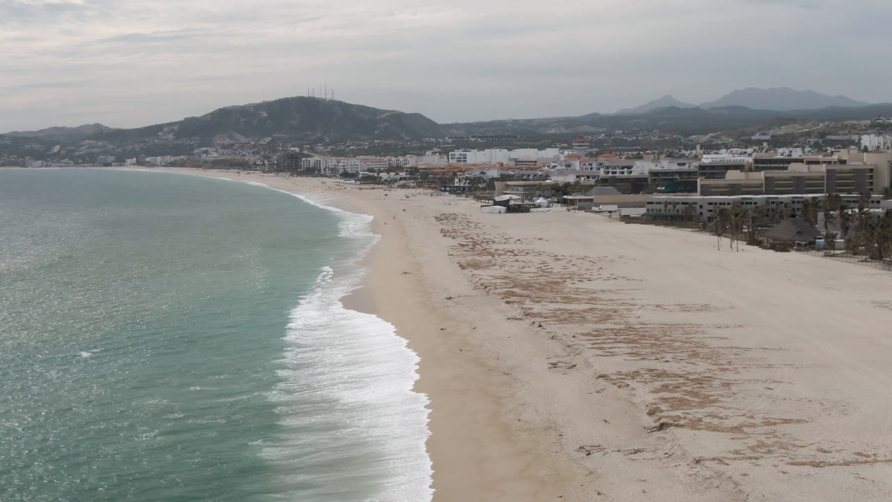 Wide aerial frame over San Jose del Cabo beach with green-blue water on the left, pale sand on the right, low buildings beyond the shore, distant mountains, and no visible people.