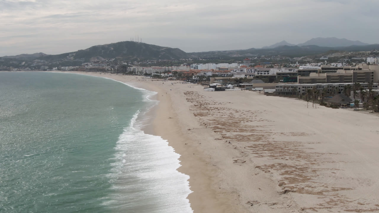 Aerial view of the San Jose del Cabo shoreline with a bright white wave band closer to the center, broad sand to the right, low urban buildings inland, mountains in haze, and no visible people.