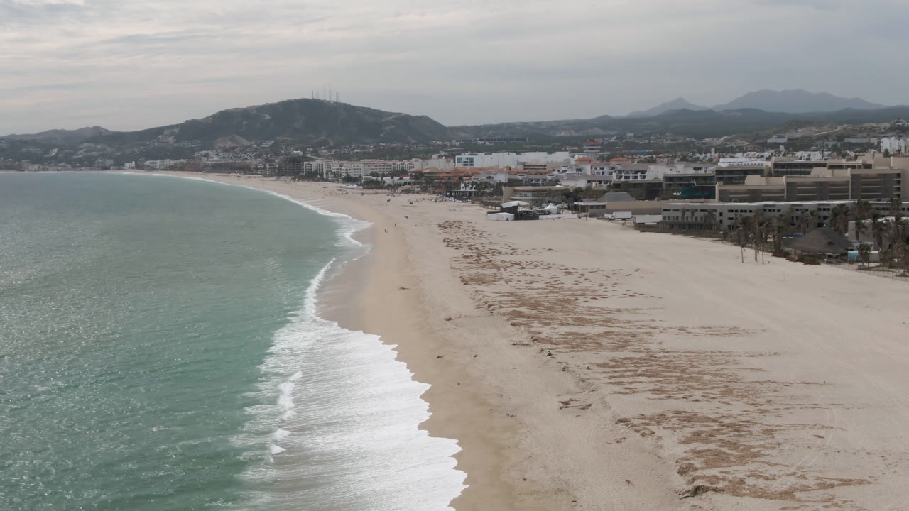 High view of a beach with turquoise water, white surf, sand, buildings, and mountains.