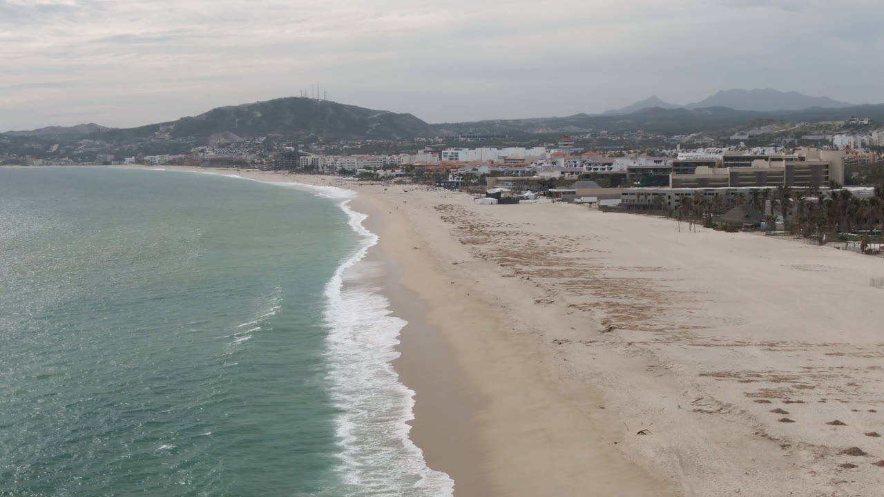 Aerial view of a pale beach, green-blue ocean, coastal buildings, and mountains