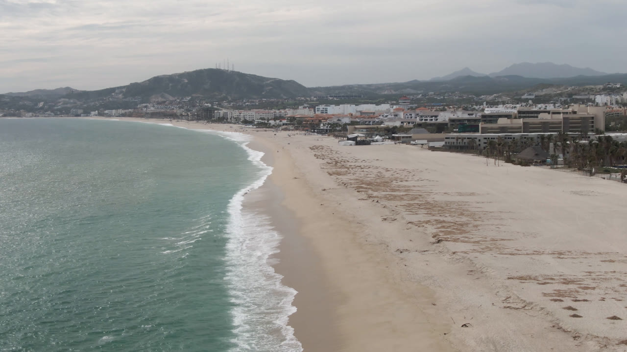 Aerial view one second into the San Jose del Cabo clip, with the surf line slightly farther along the beach, low buildings to the right, hazy hills beyond, and no visible people.