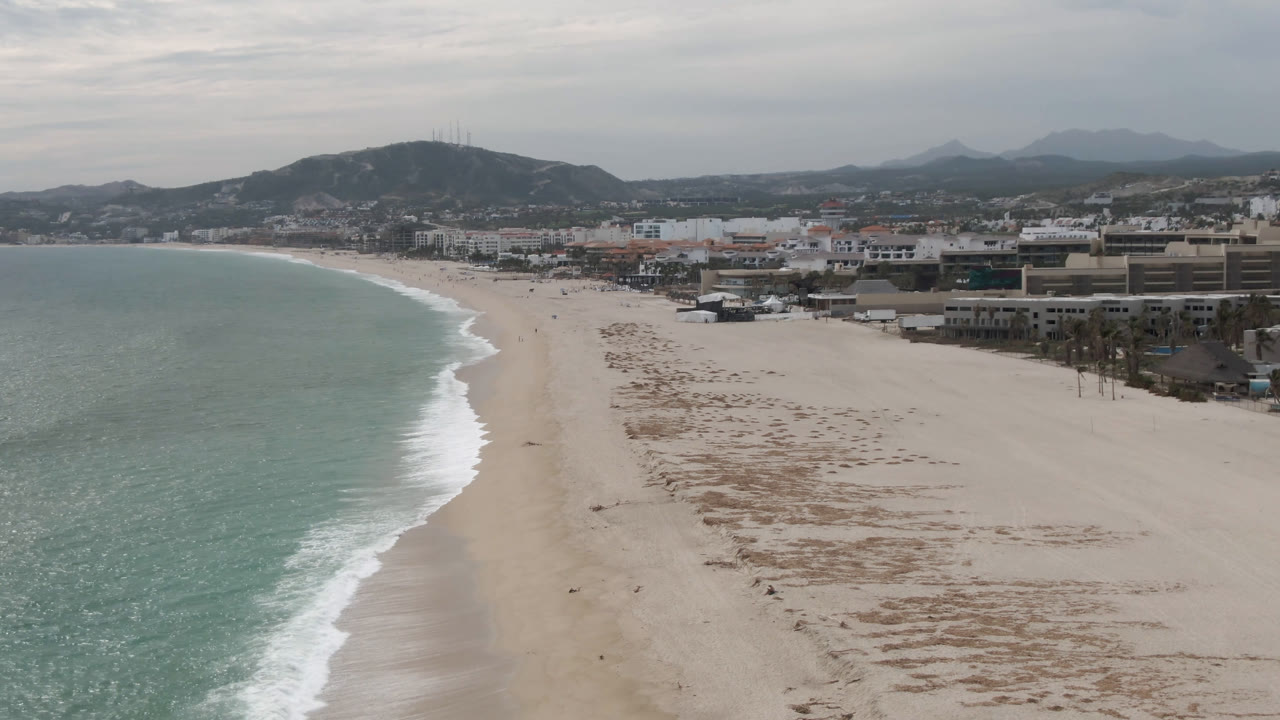 Wide aerial frame over San Jose del Cabo with pale sand in the foreground, green-blue surf on the left, low beachfront buildings beyond the shore, and hazy hills under a cloudy sky.