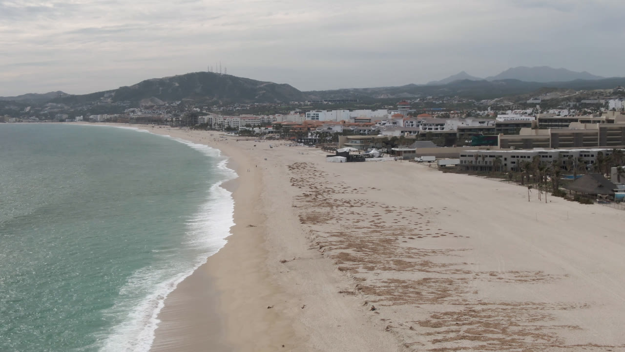 San Jose del Cabo shoreline from above, with the surf line curving deeper into frame, a broad sandy beach, scattered low buildings, and hills stretching across the horizon.