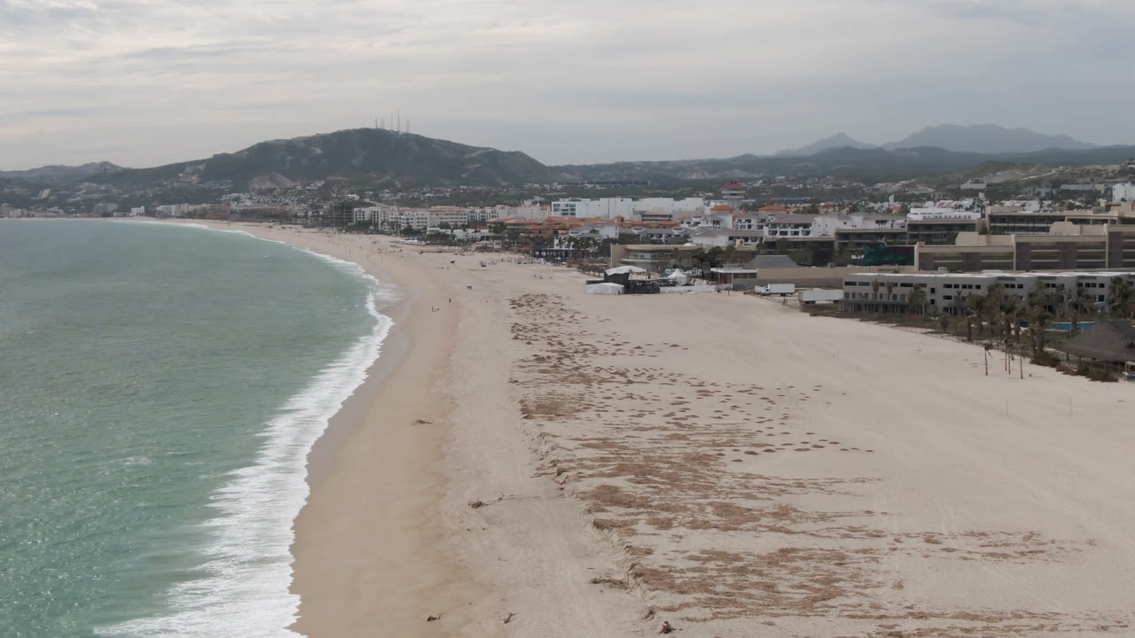 Aerial view over San Jose del Cabo beach with green-blue ocean on the left, broad pale sand in the center, shoreline buildings on the right, hazy hills beyond, and no visible people.