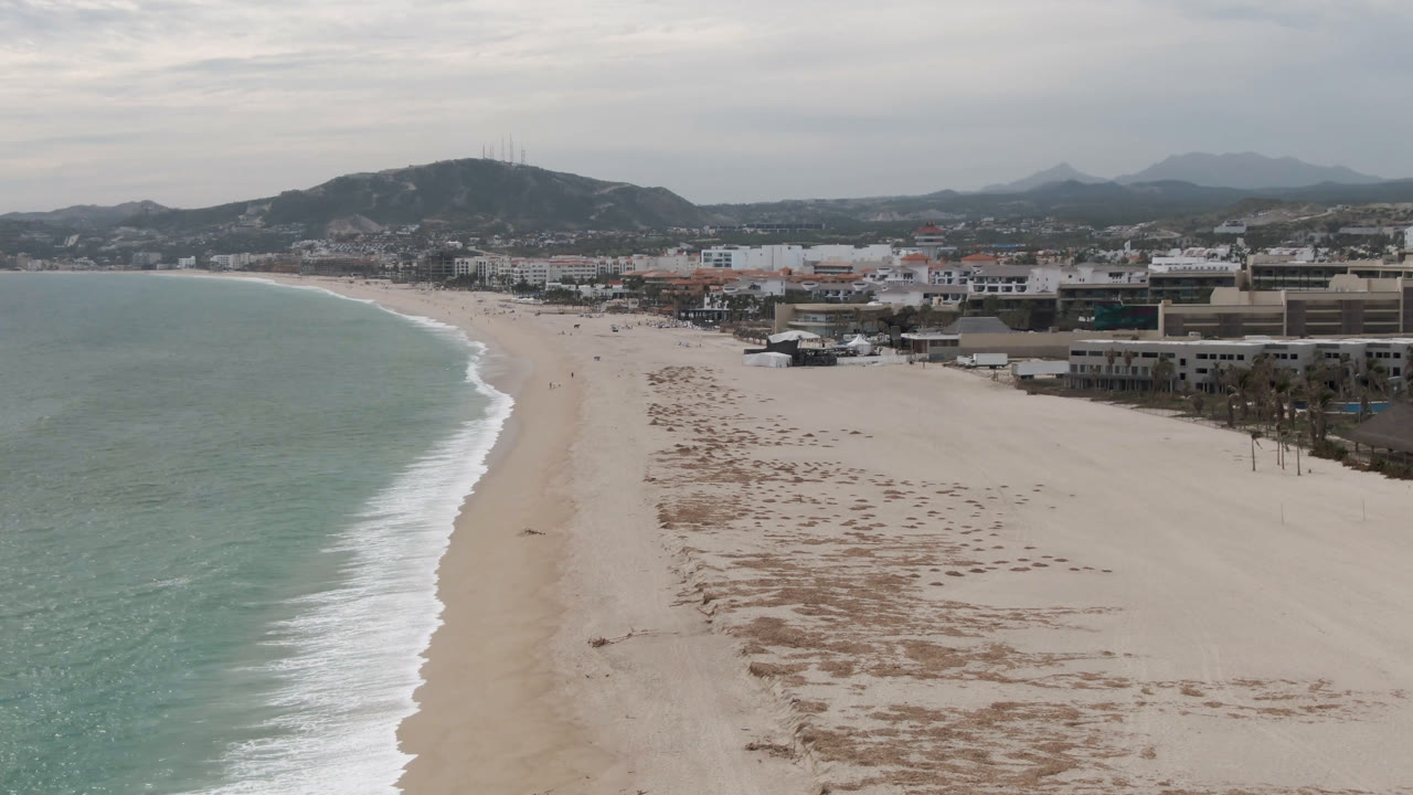Wide aerial frame of San Jose del Cabo shoreline with the surf line curving farther into the beach, open sand stretching toward low buildings, distant mountains, and no visible people.