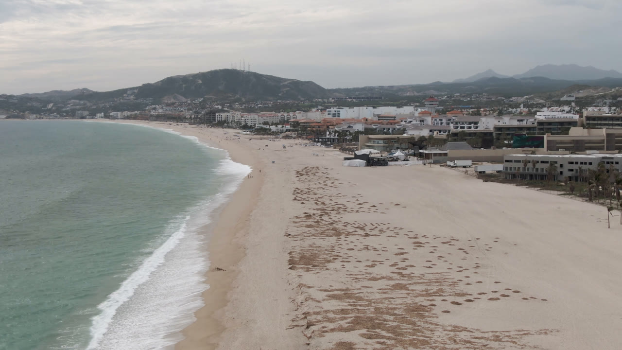 Aerial view over San Jose del Cabo beach with pale green ocean on the left, wide sand in the center, low buildings to the right, distant hills, and no visible people.