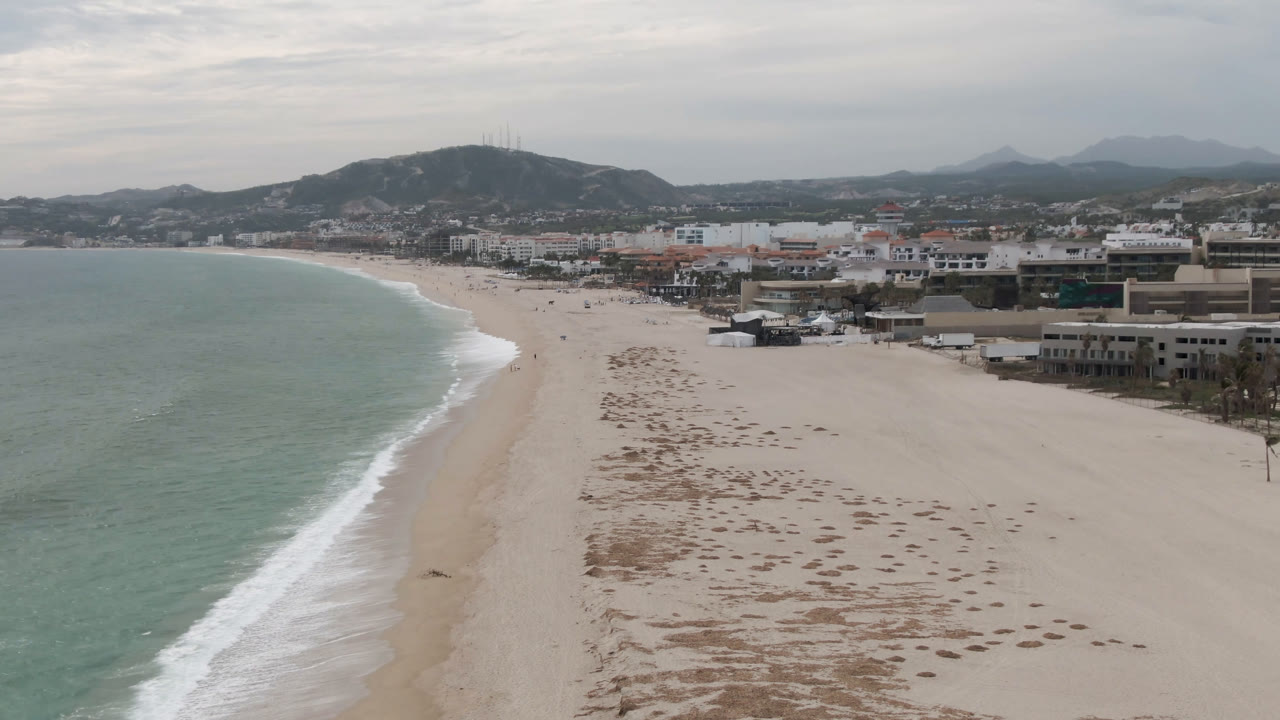San Jose del Cabo shoreline from above, with the curved surf line closer to the center, empty-looking sand, beachfront buildings, hazy hills, and no visible people.