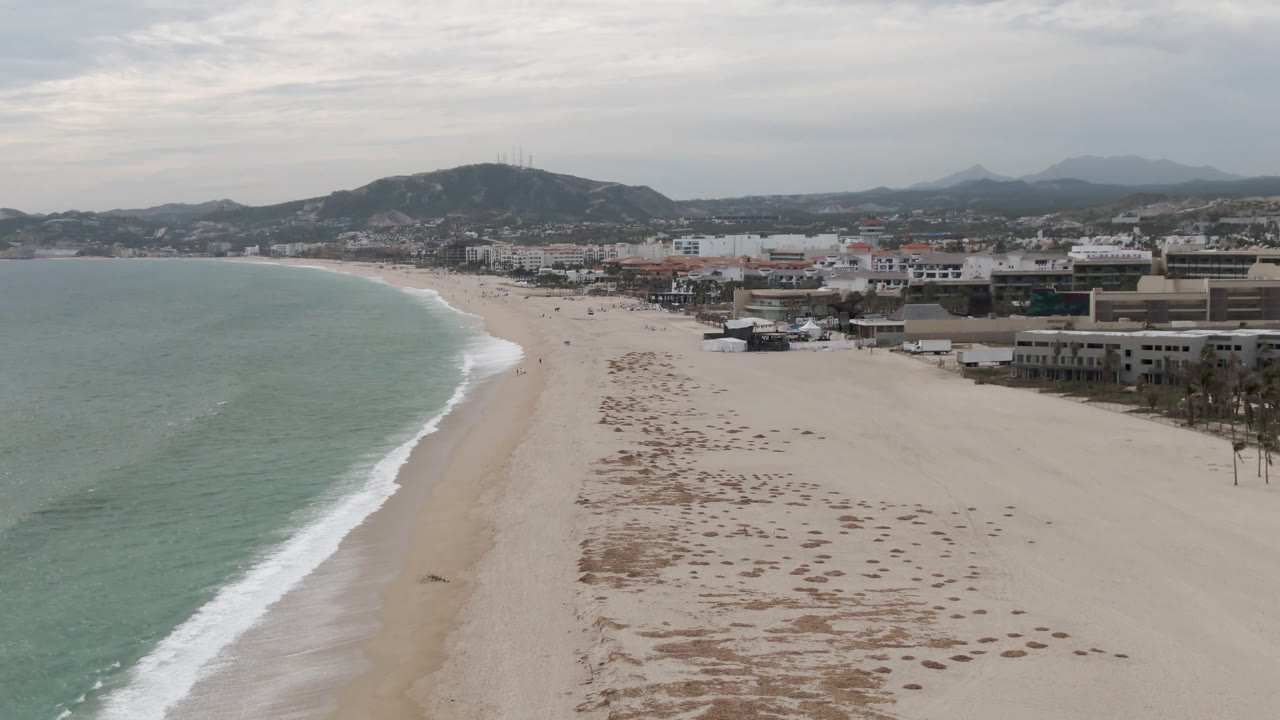 Aerial beach view with green water, white surf, broad sand, town buildings, and mountains.