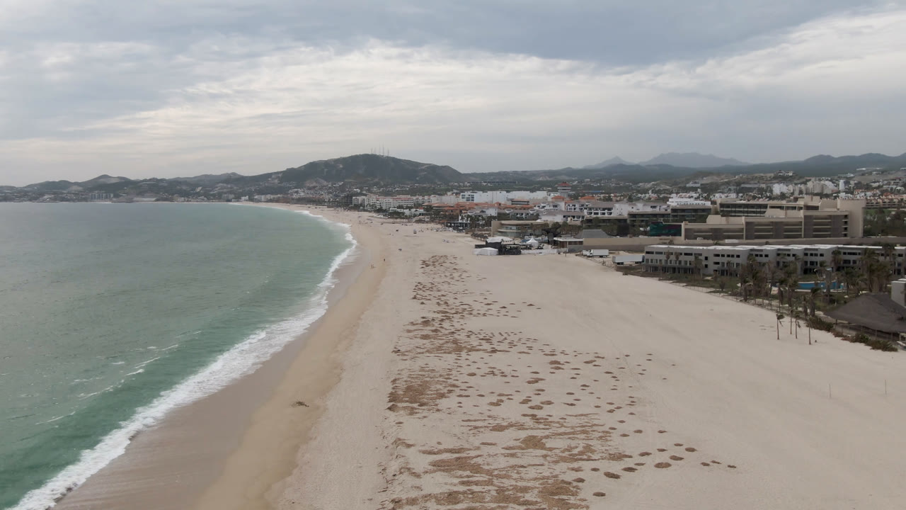 Opening aerial frame over San Jose del Cabo beach, with green-gray ocean on the left, pale sand in the center, beachfront buildings on the right, low hills beyond, and no visible people.