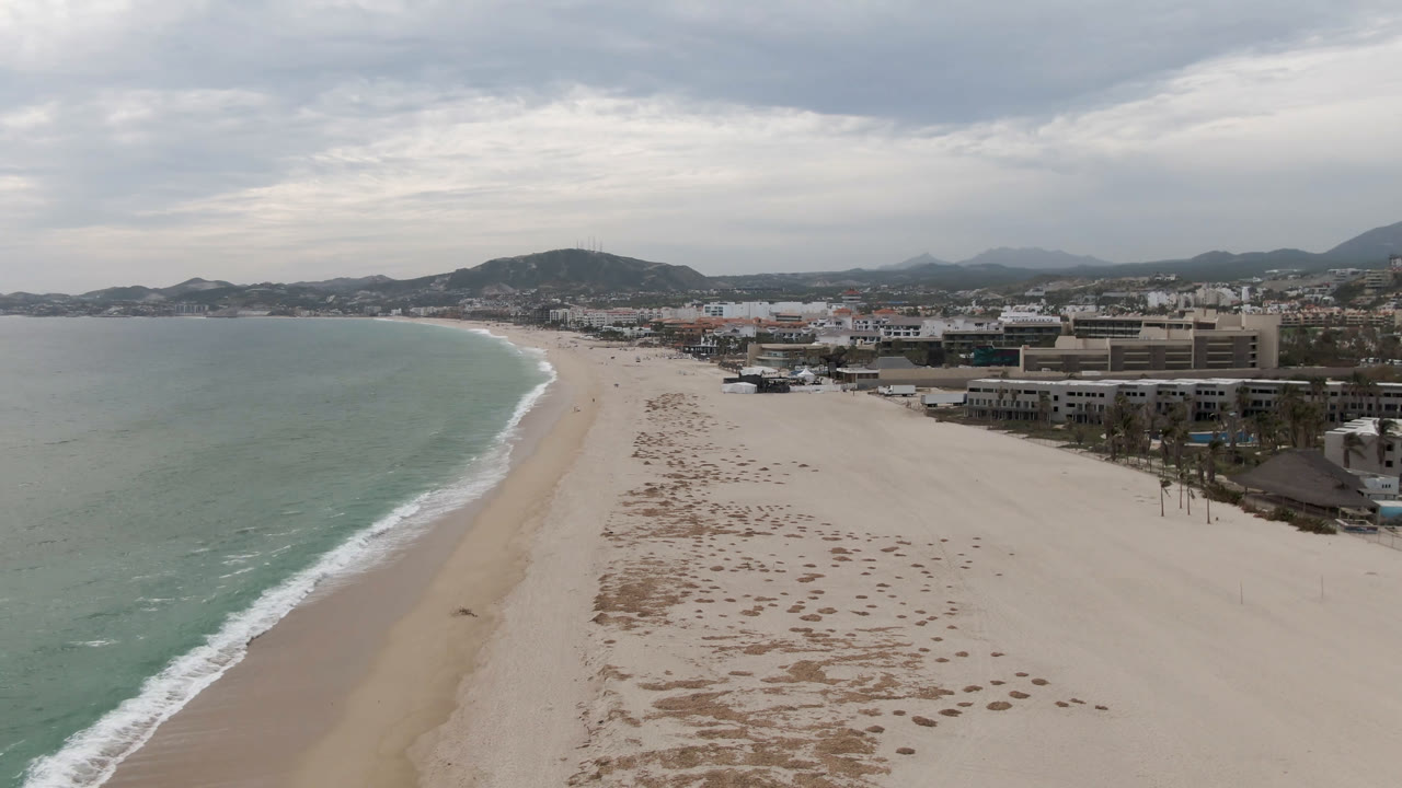 One second into the San Jose del Cabo aerial, the surf line runs diagonally along the wide beach, with town buildings and hills under layered clouds, and no visible people.