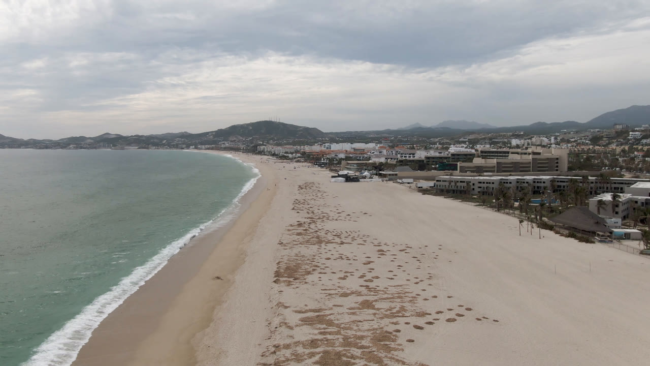 Aerial coastline view with ocean, empty beach, buildings, hills, and cloudy sky.