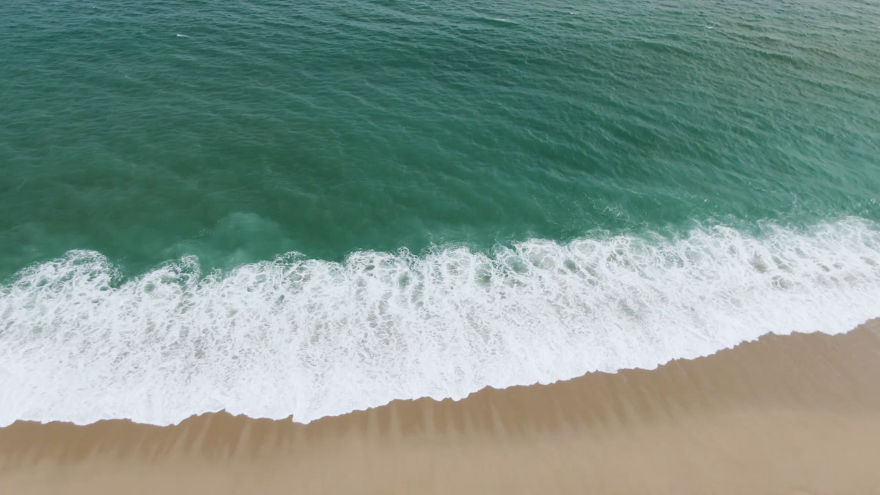 Overhead view of green-blue water and white surf reaching tan sand in San Jose del Cabo, with no visible people.