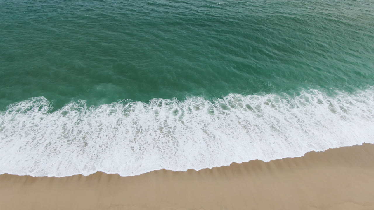 A foamy wave line spreads wider across the sandy San Jose del Cabo shore, leaving pale wash marks and no visible people.