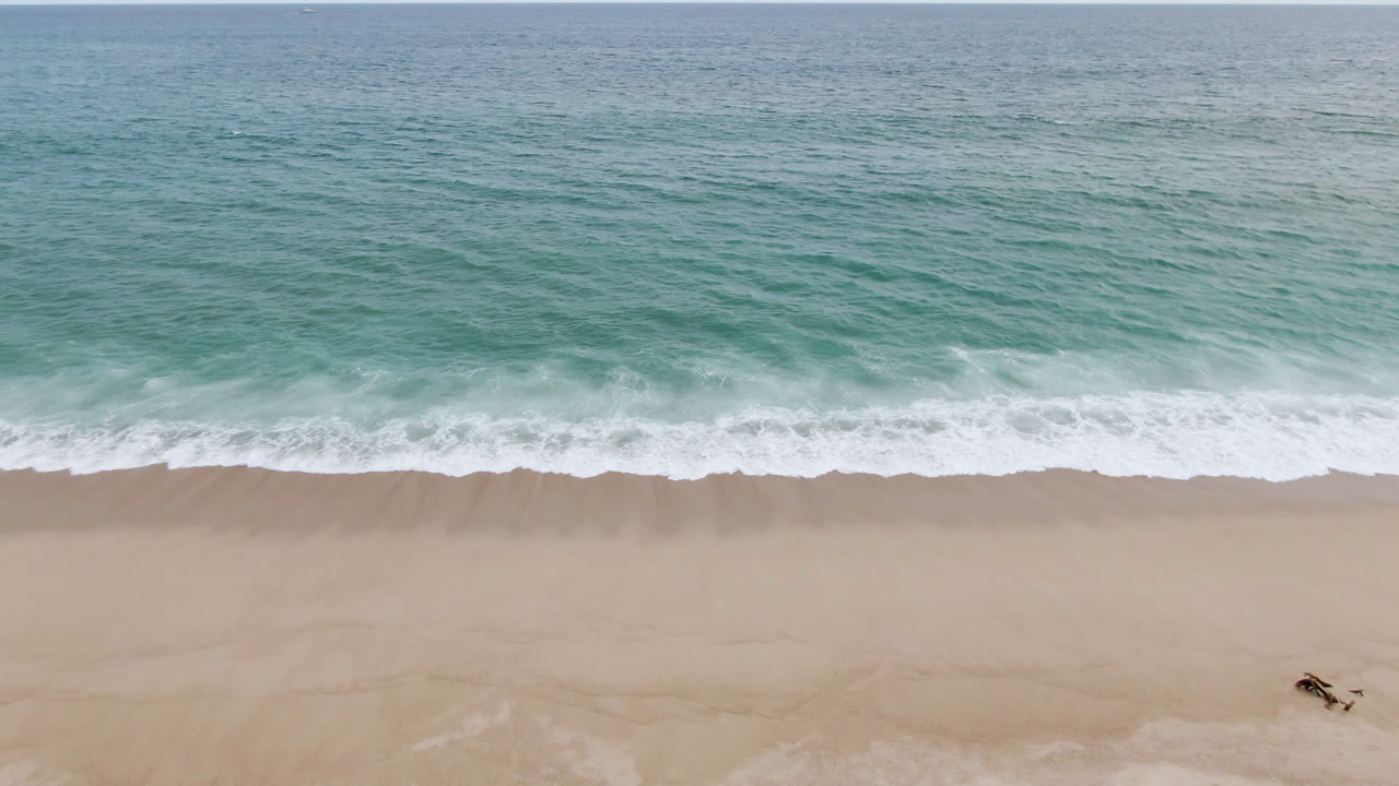 Aerial view of the San Jose del Cabo shoreline with teal ocean, white surf, tan sand, and no visible people in this opening frame.