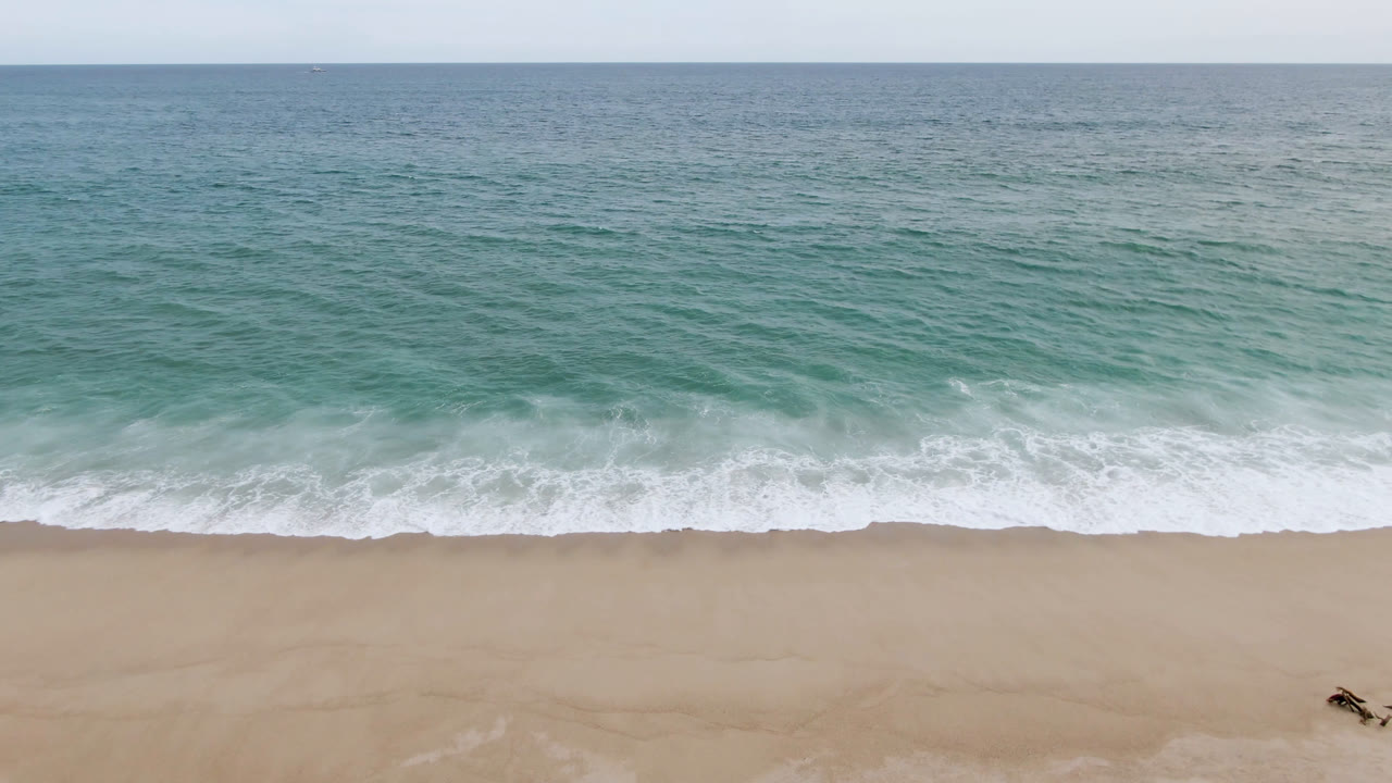 Aerial San Jose del Cabo beach frame with rolling surf at mid-frame and a small distant person with a surfboard near the lower right sand.