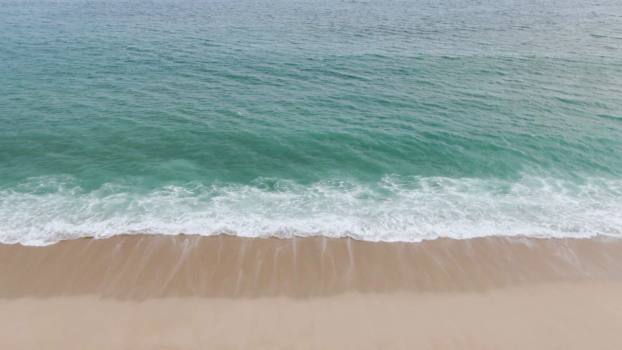 A San Jose del Cabo aerial frame shows the wave edge pushed slightly farther onto the wet sand beneath textured green-blue water, with no visible people.
