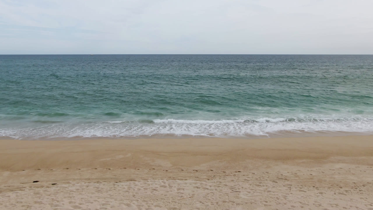 Wide San Jose del Cabo beach view with pale sand, teal ocean, a level horizon, thin clouds, and no visible people at the start of the clip.