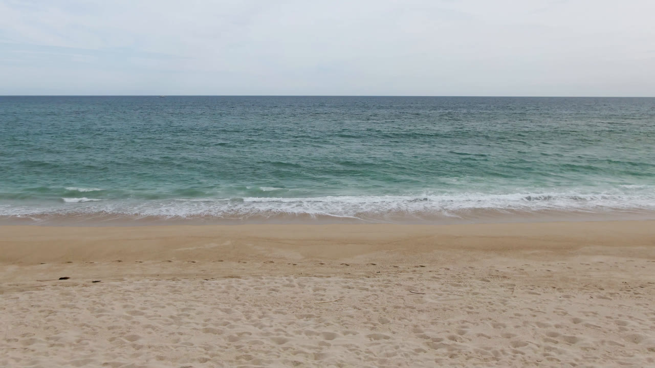 San Jose del Cabo shoreline frame with textured sand in front, a narrow band of white surf, open ocean beyond, and no visible people.