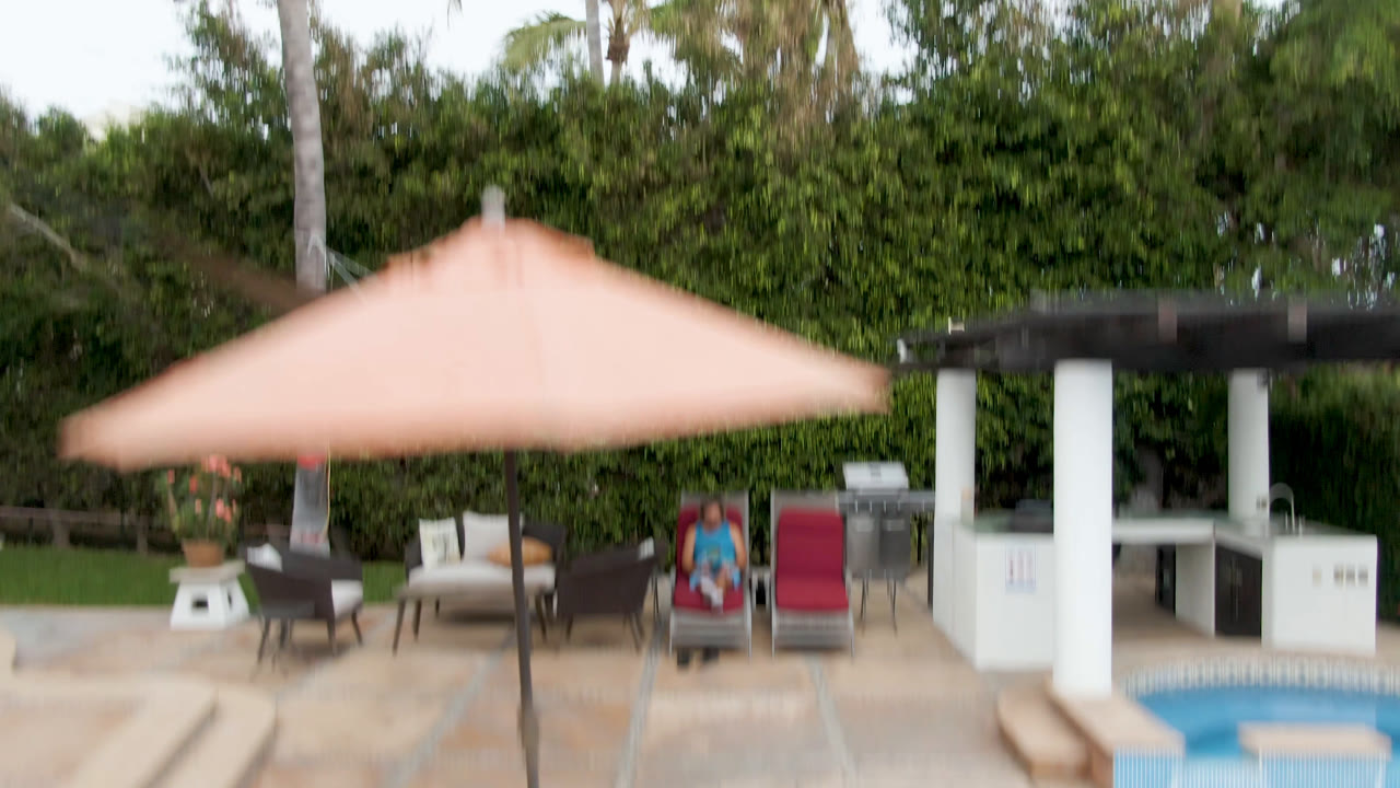Elevated view of a pool patio in San Jose del Cabo with a peach umbrella, lounge chairs, palms, hedges, and possible people or human imagery near the seating area.