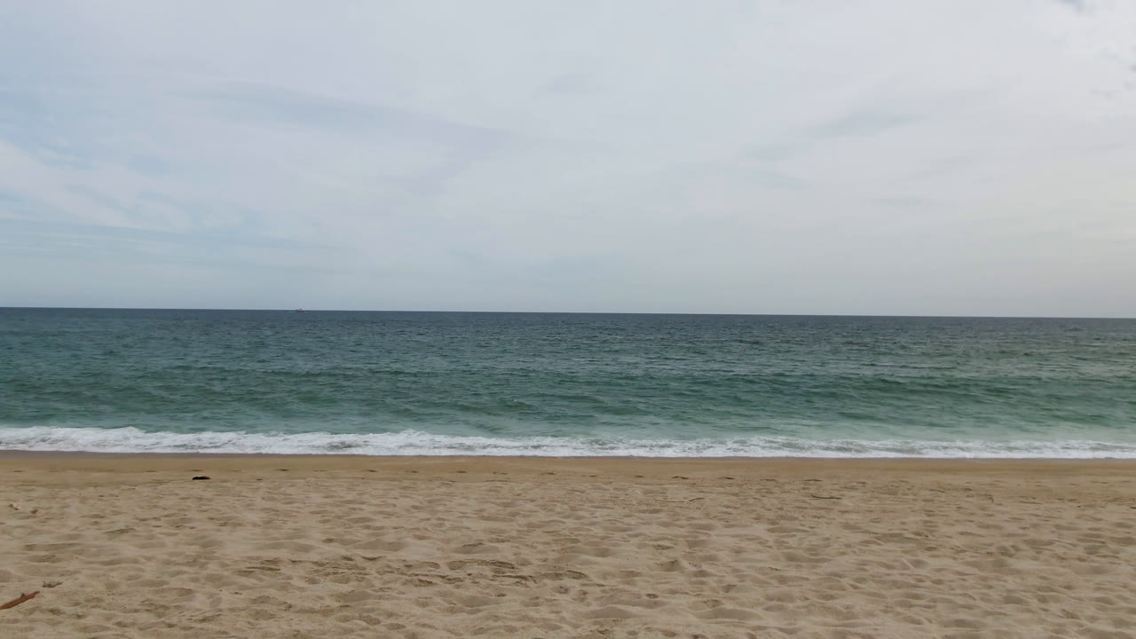 Wide San Jose del Cabo beach frame with empty white sand in the foreground, low surf beyond it, a straight ocean horizon, pale clouds overhead, and no visible people.
