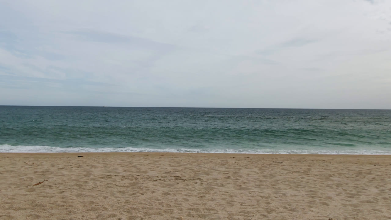 San Jose del Cabo shoreline frame showing open sand, green-blue water, a thin white wave line, soft cloud cover, and no visible people.