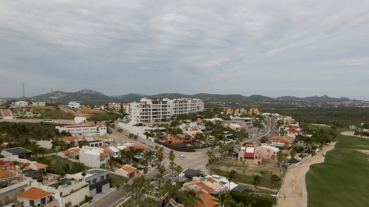 Wide aerial frame over San Jose del Cabo with green fairway at right, red-roofed homes in the foreground, white apartment buildings near center, low hills behind, and no visible people.