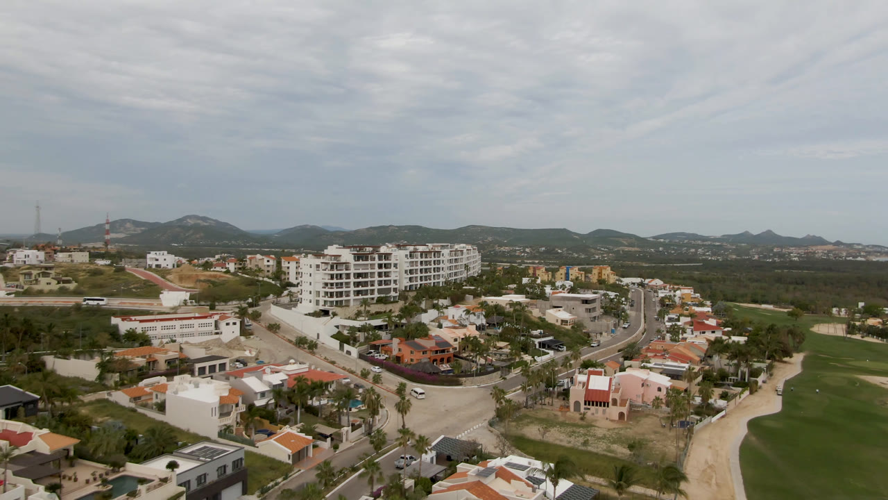 Aerial view in San Jose del Cabo with a curving road leading past white mid-rise buildings, a golf fairway and sand area at left, houses in the lower frame, and no visible people.