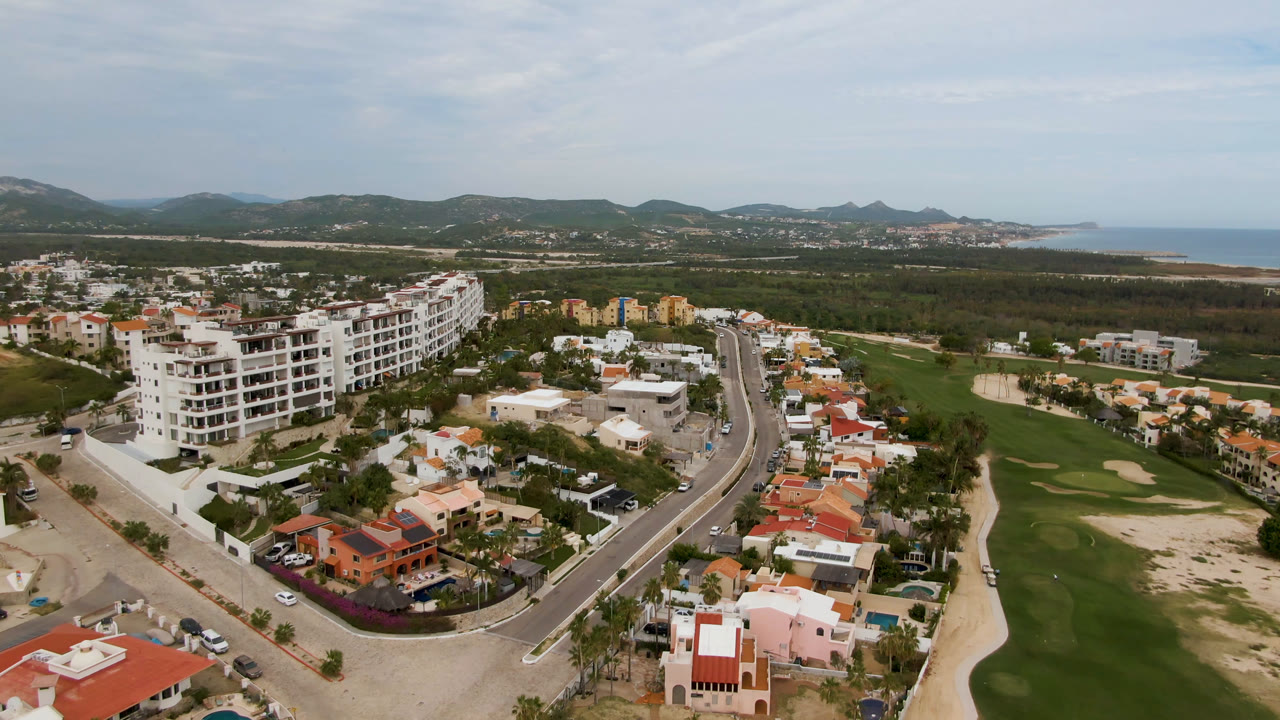 San Jose del Cabo aerial frame with white apartment buildings left of center, red-roofed homes in the foreground, green fairways and sand traps to the right, distant hills and sea, and no visible people.