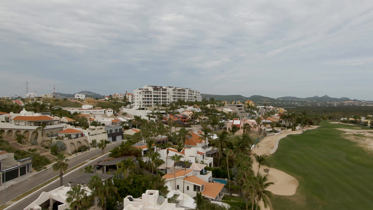 Aerial view in San Jose del Cabo with the golf course centered, sand traps along the edge, white buildings beyond the road, distant hills, and no visible people.