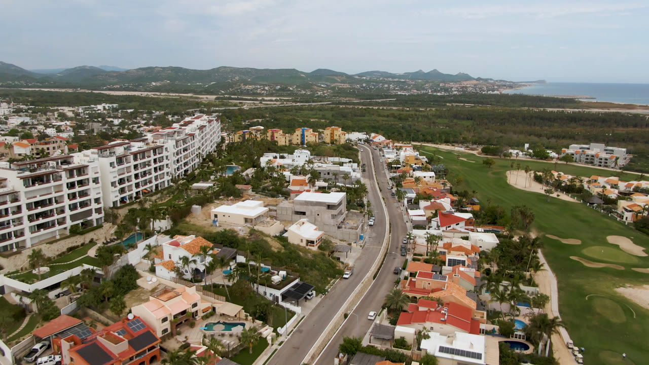 The San Jose del Cabo aerial angle continues with apartment blocks closer to the left, villas below, a paved road through the center, golf grass to the right, and no visible people.
