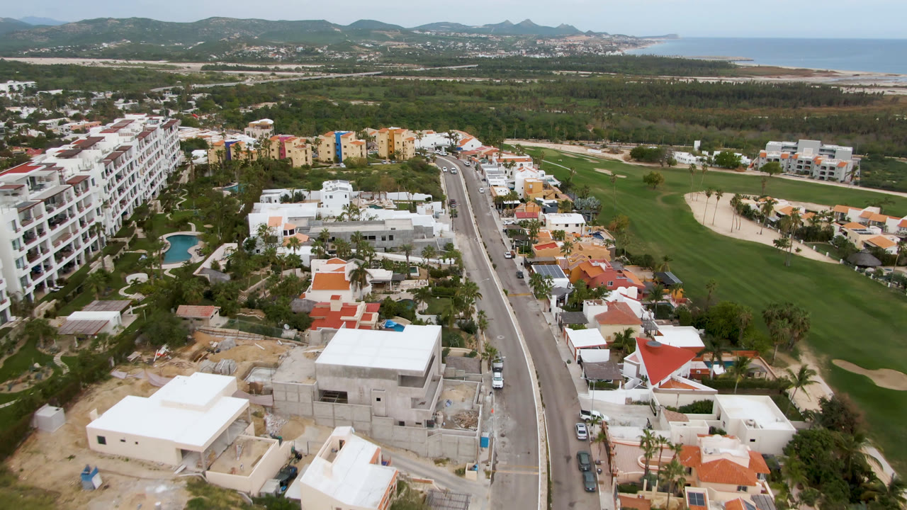 San Jose del Cabo aerial view with the road centered between villas and apartment blocks, golf course greens to the right, coastline on the horizon, and no visible people.