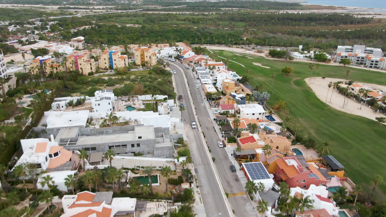 San Jose del Cabo aerial view with colorful apartment buildings, villas with pools, cars on the road, golf course grass, sand areas, and no visible people.