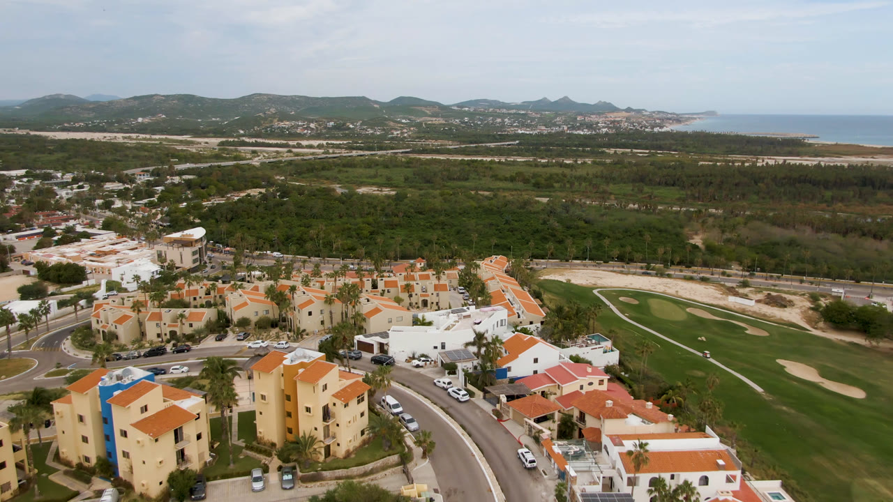 Wide aerial frame over San Jose del Cabo with orange-roofed homes in front, golf course fairways to the right, green tree cover, low hills, distant sea, and no visible people.