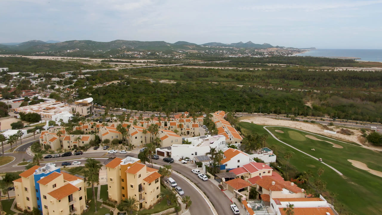 San Jose del Cabo aerial view with foreground apartments and villas, a curved road with cars, golf course grass and bunkers, thick greenery, hills, coastline, and no visible people.