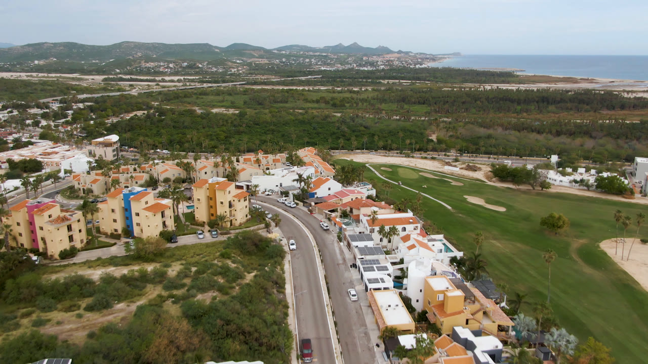 Aerial view in San Jose del Cabo with apartment-style buildings and villas in the foreground, golf greens to the right, coastal water on the horizon, and no visible people.