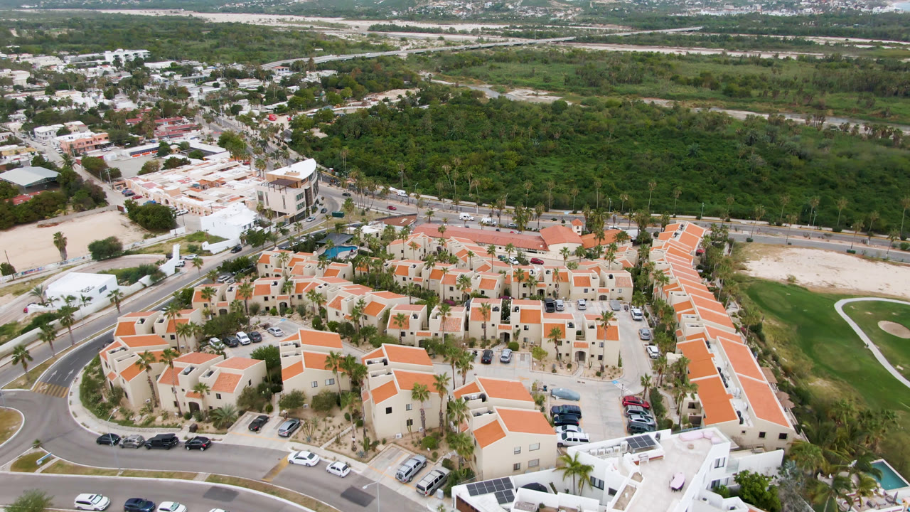 Aerial frame over San Jose del Cabo showing clustered orange-roofed residences, parked cars, curved roads, palms, and a broad green area beyond, with no visible people.