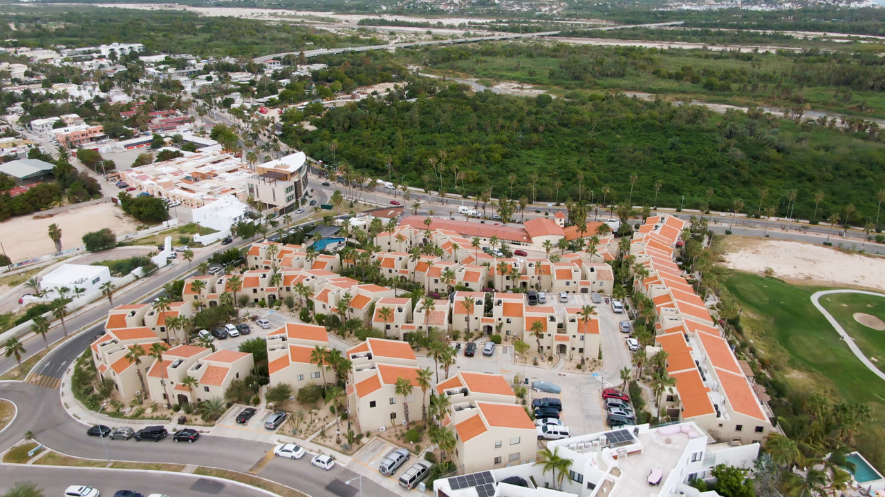 High aerial view in San Jose del Cabo with the residential complex centered, a white building at lower right, parked cars along the road, and dense green landscape behind, with no visible people.