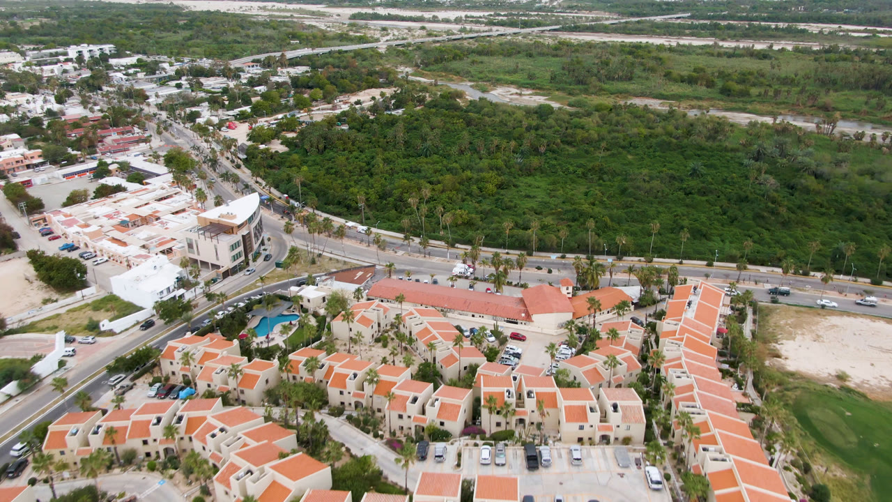 Second aerial frame in San Jose del Cabo, Mexico, showing the main road more centrally, small vehicles along the lanes, orange rooftops below, dense green vegetation behind, and no visible people.