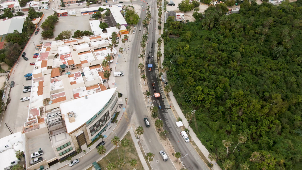 Aerial frame in San Jose del Cabo, Mexico, with the road more centered between the white building, parked cars, palms, thick vegetation, and no visible people.