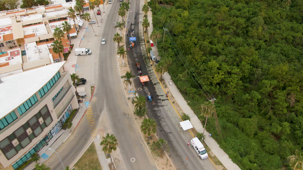 Aerial frame from San Jose del Cabo showing a curved road beside a beige commercial building, palm trees, parked vehicles, dense green vegetation, and no visible people.