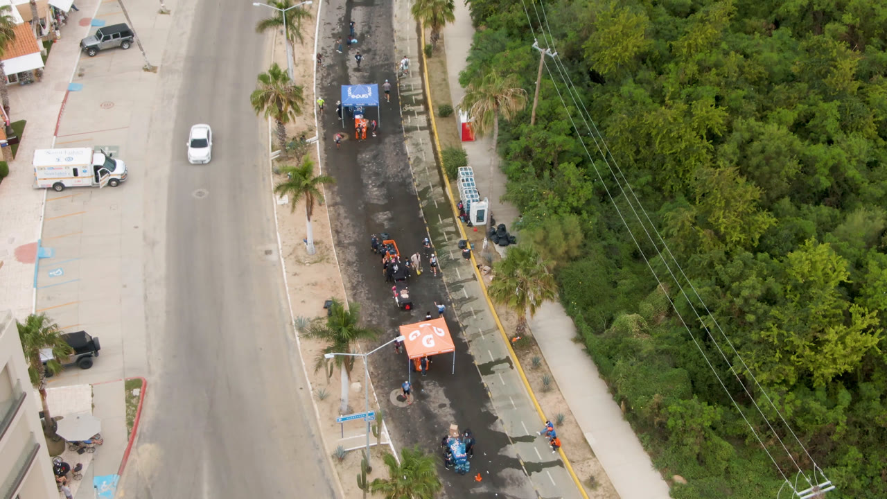Overhead frame in San Jose Del Cabo showing a road beside a narrow race lane with small runners, orange and blue tents, parked vehicles, palm trees, and dense green vegetation.