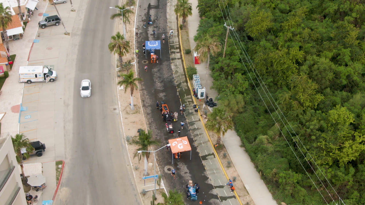 Aerial frame in San Jose Del Cabo with a white car on the main road, small runners clustered along the event lane, service tents, palm trees, sidewalks, and thick roadside vegetation.