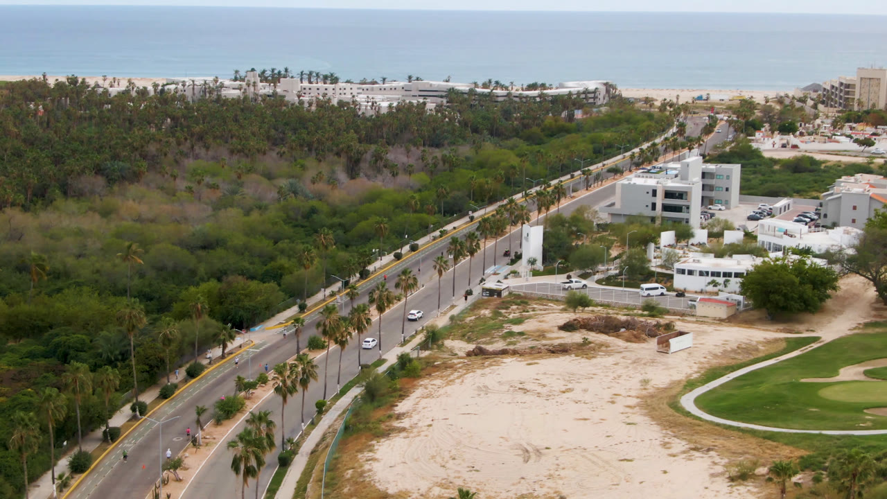 Opening aerial frame over San Jose del Cabo, Baja California Sur, with a palm-lined road at left, sandy foreground, curved golf greens at right, low buildings near center, beach hotels, ocean horizon, and no visible people.