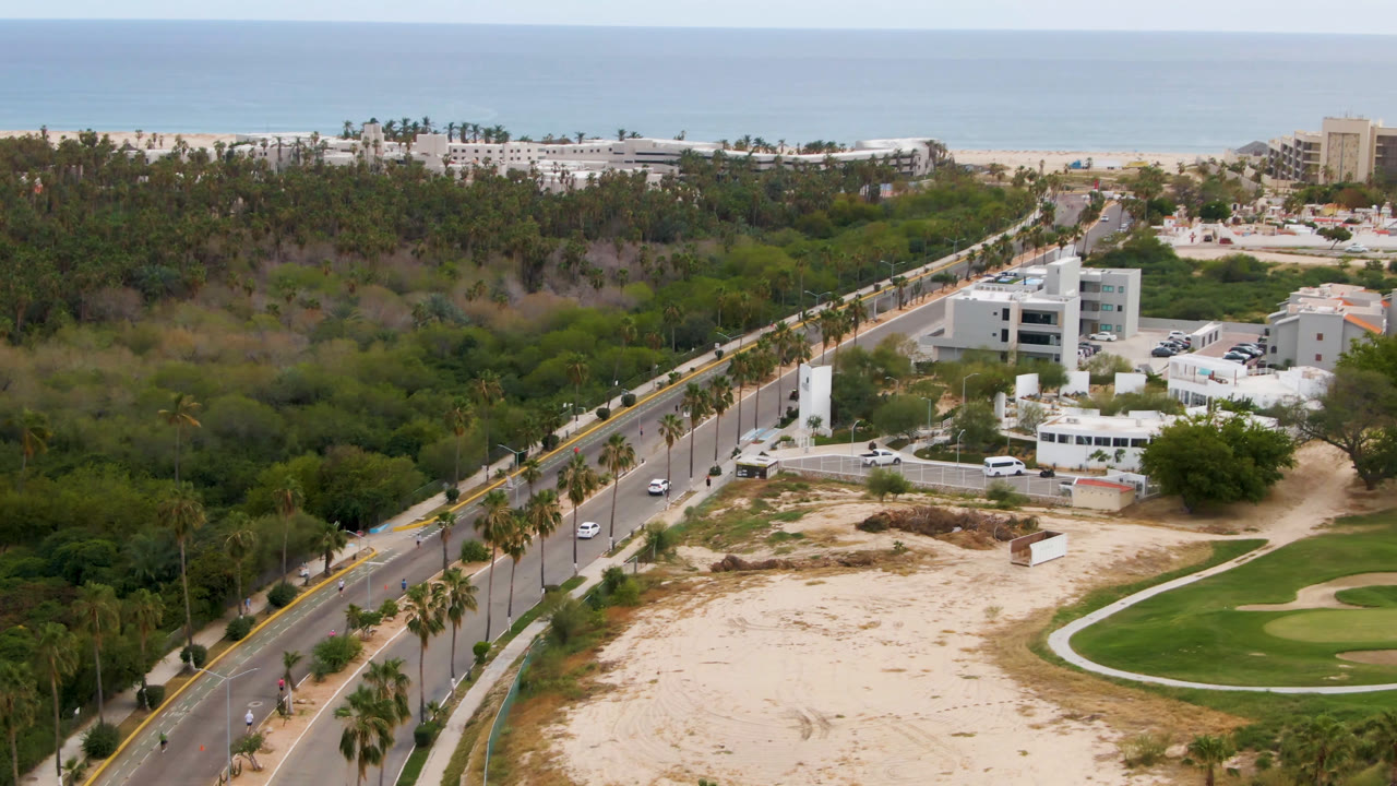 One second into the clip, the San Jose del Cabo aerial view keeps the road and palms along the left diagonal, with white buildings in the midground, golf course curves at lower right, distant beach structures, ocean, and no visible people.